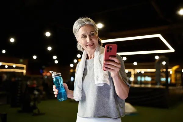 Cheerful senior gray haired sportswoman in cozy attire looking at phone with water bottle in hands — Stock Photo