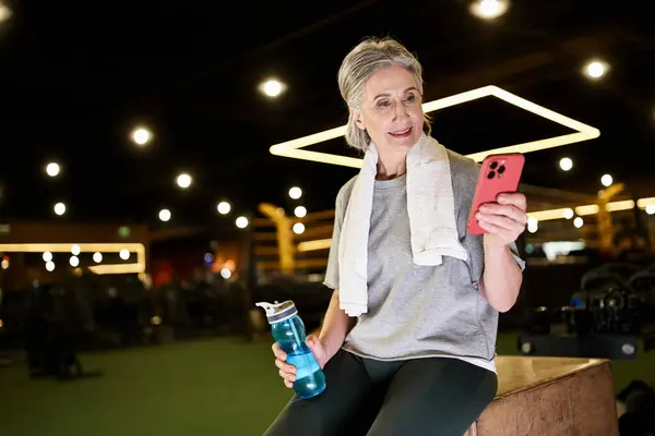 Gioiosa donna dai capelli grigi matura sportiva in abbigliamento comodo guardando il telefono con bottiglia d'acqua in mano — Foto stock