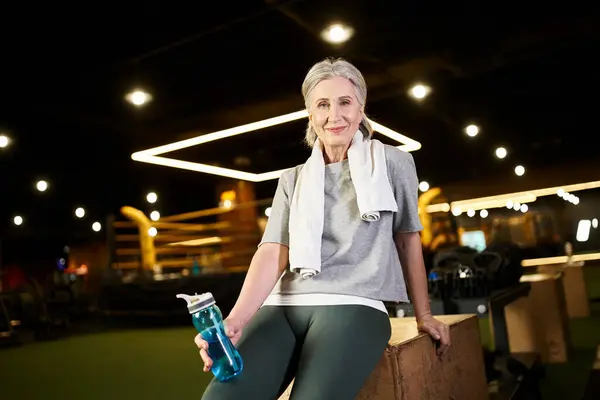 Веселая пожилая женщина с полотенцем и бутылкой воды улыбаясь в камеру во время отдыха после тренировки — стоковое фото