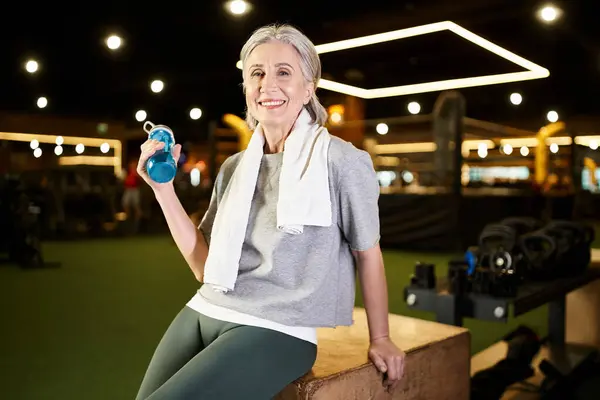 Довольно веселая зрелая женщина с полотенцем и бутылкой улыбаясь в камеру во время отдыха после тренировки — стоковое фото
