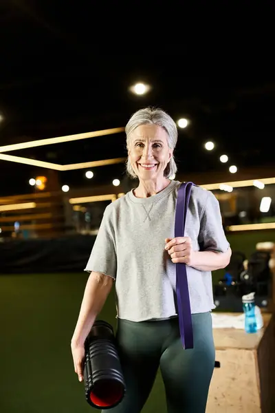 Cheerful mature sportswoman posing with fitness expander and weight bag and smiling at camera — Stock Photo