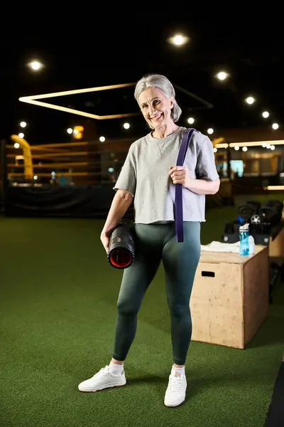 Весело зрелая женщина в спортивной одежде позирует с фитнес-экспандером и мешком веса и улыбается в камеру — стоковое фото