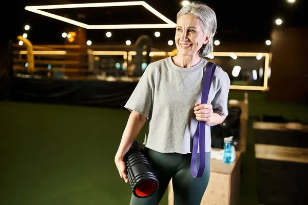 Jolly good looking mature woman posing in gym with fitness expander and weight bag and looking away — Stock Photo