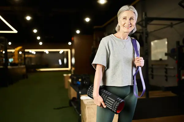 Atractiva mujer mayor alegre posando en el gimnasio con expansor de fitness y bolsa de peso y mirando hacia otro lado - foto de stock