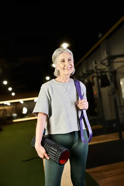 Fröhlich attraktive reife Frau posiert im Fitnessstudio mit Fitness-Expander und Gewichtsbeutel und schaut weg — Stockfoto
