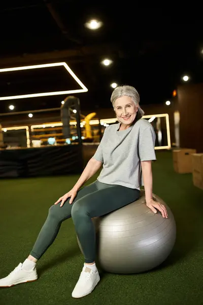 Радостная зрелая спортсменка в уютном наряде сидит на фитнес-мяче и улыбается перед камерой в тренажерном зале — стоковое фото