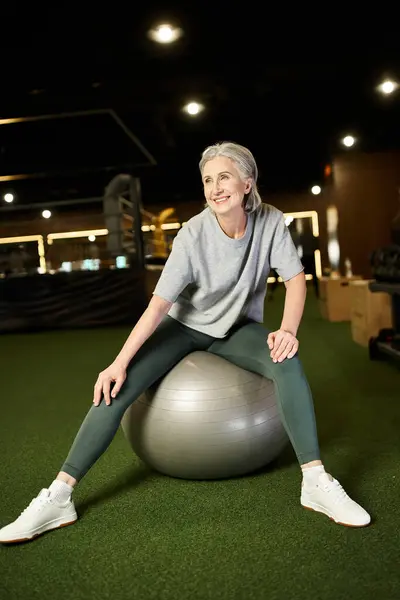 Красивая веселая зрелая женщина в спортивной одежде сидит на фитнес-мяч и смотрит в сторону в то время как в тренажерном зале — стоковое фото