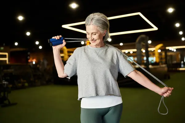 Joyeuse mature belle femme en vêtements de sport posant avec corde à sauter dans la salle de gym et regardant loin — Photo de stock