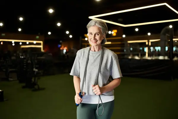 Gaie femme âgée en tenue de sport posant avec corde sautante et souriant à la caméra dans la salle de gym — Photo de stock
