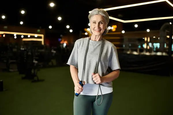 Joyous mature sportswoman in cozy attire posing with skipping rope and smiling at camera in gym — Stock Photo
