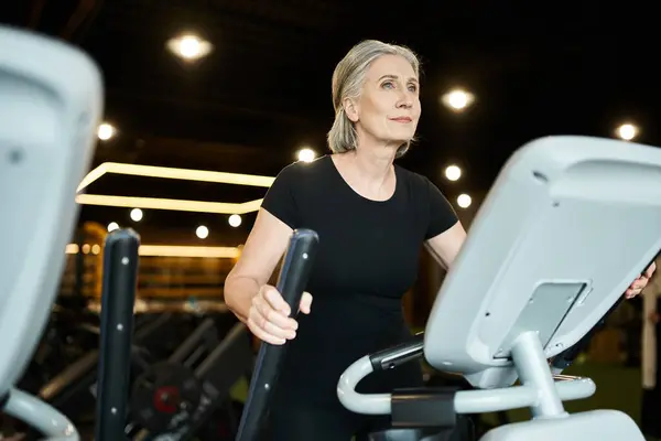 Good looking jolly mature sportswoman in black t shirt exercising actively on elliptical trainer — Stock Photo
