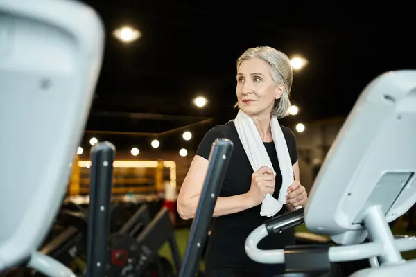 Jolly athletic senior woman exercising on cross trainer while in gym with towel on shoulders — Stock Photo
