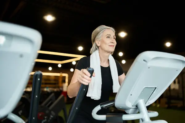 Mujer mayor deportiva positiva haciendo ejercicio en cross trainer mientras está en el gimnasio con toalla en los hombros - foto de stock