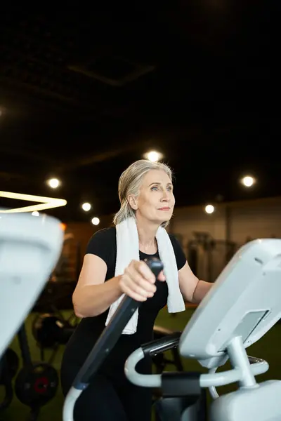 Beautiful athletic mature woman exercising on cross trainer while in gym with towel on shoulders — Stock Photo