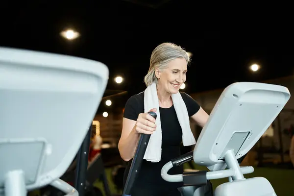 Sporty mature cheerful woman with gray hair and towel on shoulders exercising on cross trainer — Stock Photo