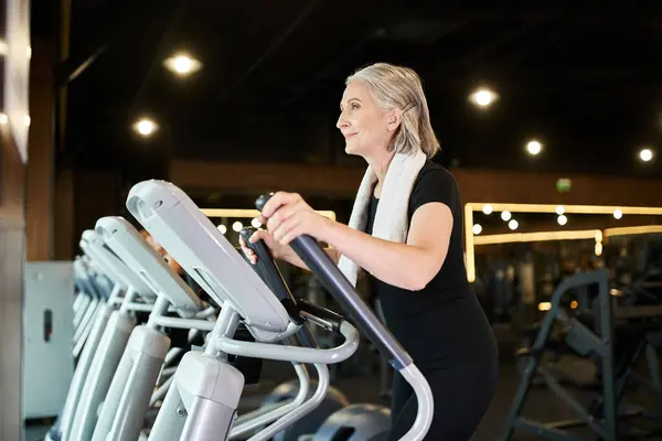 Appealing senior cheerful sportswoman with towel on shoulders exercising on elliptical trainer — Stock Photo