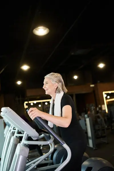 Cheerful sporty mature woman exercising on cross trainer while in gym with towel on shoulders — Stock Photo
