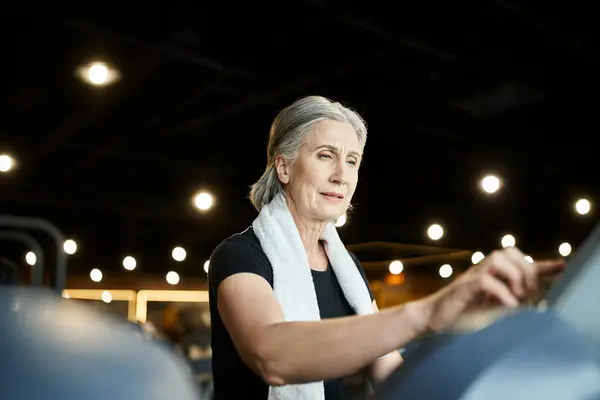 Athletic jolly senior woman in sporty attire with towel on shoulders running on treadmill in gym — Stock Photo