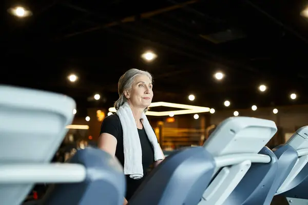 Appealing mature sportswoman in black t shirt with towel on shoulders training on treadmill actively — Stock Photo