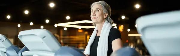 Sporty senior woman in sporty attire with towel on shoulders running on treadmill in gym, banner — Stock Photo
