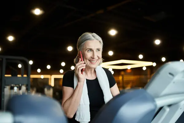 Beautiful cheerful senior woman with towel on shoulders talking by phone while on treadmill — Stock Photo