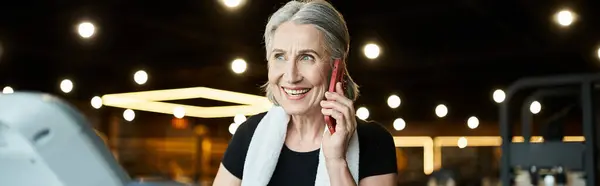 Positive senior woman with towel on shoulders talking by phone while on treadmill in gym, banner — Stock Photo