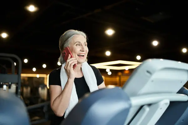 Appealing joyous mature woman with towel on shoulders talking by phone while on treadmill in gym — Stock Photo