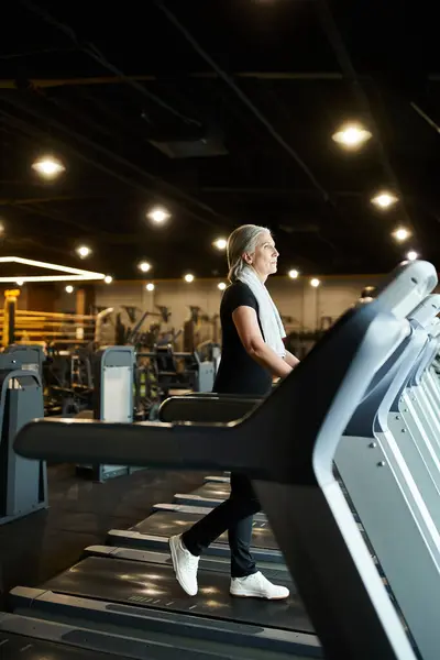 Mujer deportiva madura de buen aspecto en el entrenamiento de traje negro en la cinta de correr activamente mientras está en el gimnasio - foto de stock