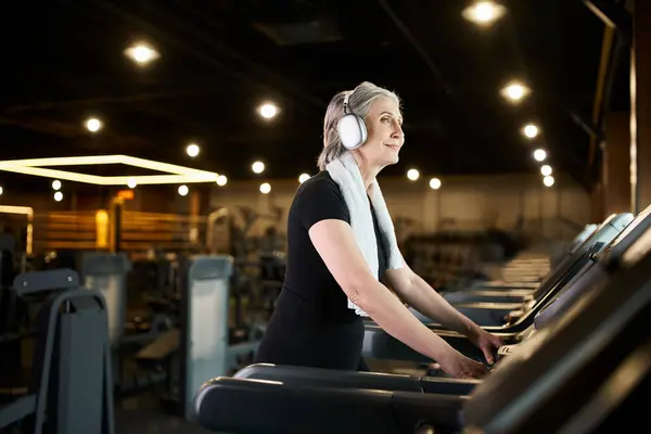 Mujer madura alegre en el entrenamiento acogedor atuendo en la cinta de correr y escuchar música en los auriculares - foto de stock