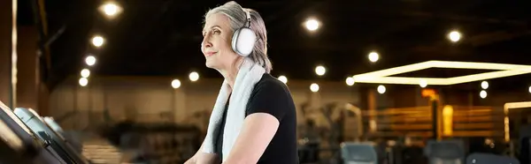 Senior femme gaie avec cheveux gris et écouteurs exercice sur tapis roulant dans la salle de gym, bannière — Photo de stock