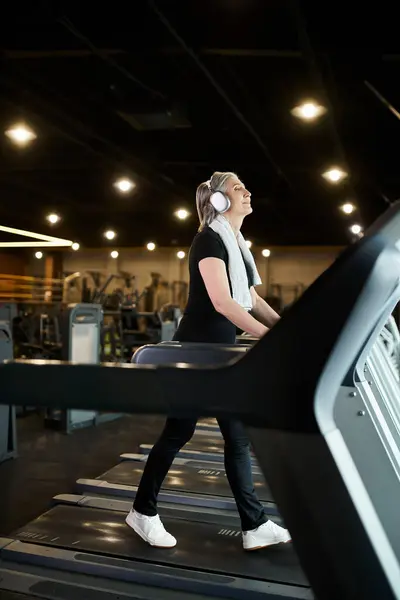 Mujer alegre madura de buen aspecto con pelo gris y auriculares haciendo ejercicio en la cinta de correr en el gimnasio - foto de stock