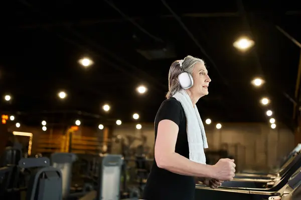Позитивная довольно зрелая женщина в уютной одежде тренировки на беговой дорожке и прослушивания музыки в наушниках — стоковое фото