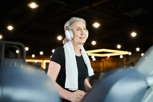 Belle femme gaie senior avec cheveux gris et écouteurs exercice sur tapis roulant dans la salle de gym — Photo de stock