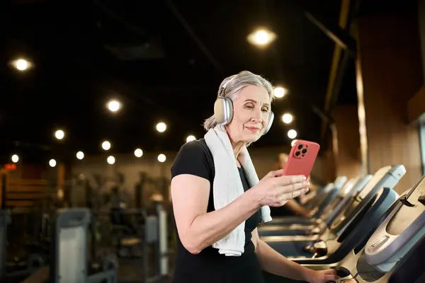 Jolly mature woman with towel on shoulders and headphones holding phone and exercising on treadmill — Stock Photo