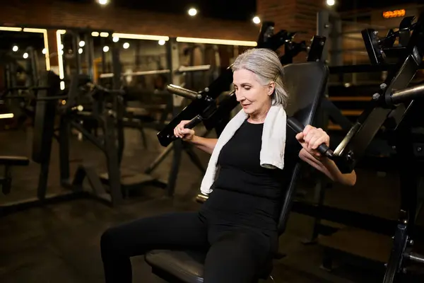 Atractiva mujer mayor en ropa deportiva acogedora con entrenamiento de pelo gris en la máquina de prensa de pecho en el gimnasio - foto de stock