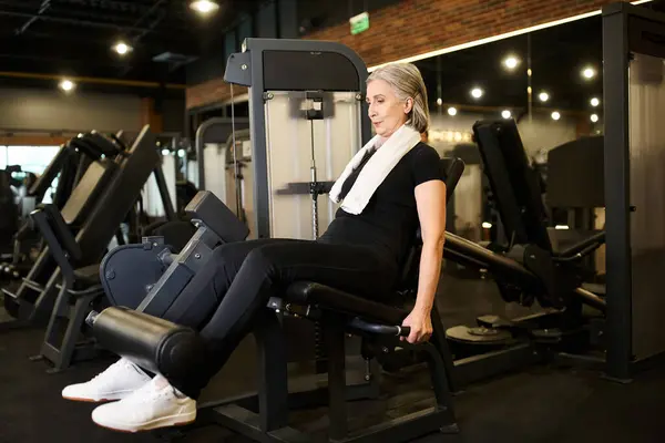 Athlétique mature femme aux cheveux gris dans l'entraînement confortable de vêtements de sport sur la machine à boucler les jambes tout en gymnase — Photo de stock