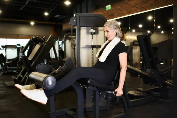 Mujer madura deportiva de buen aspecto en traje acogedor con ejercicio de toalla en la máquina de rizar la pierna en el gimnasio - foto de stock