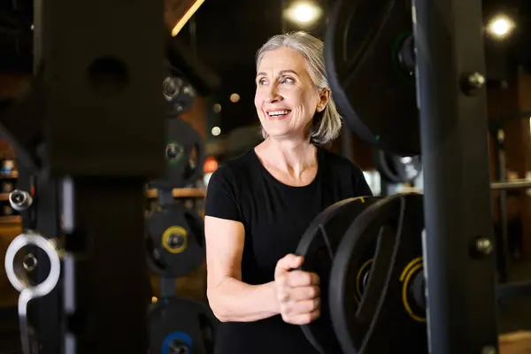 Fröhliche grauhaarige Seniorin in kuscheliger Kleidung, die im Fitnessstudio neben Hantelscheiben wegschaut — Stockfoto