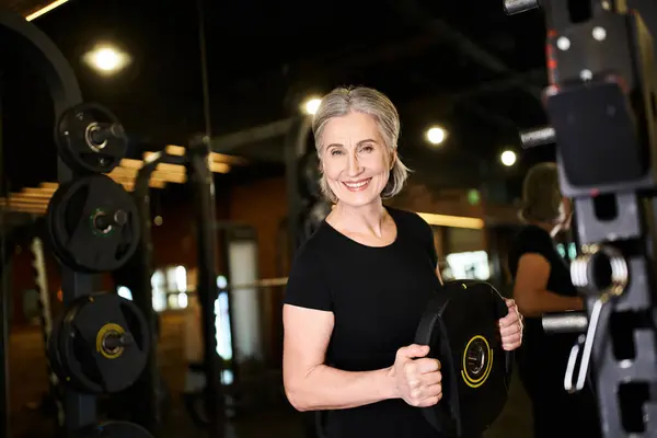 Gut aussehende, reife Frau mit grauen Haaren, die die Gewichtsscheibe hält und im Fitnessstudio in die Kamera lächelt — Stockfoto