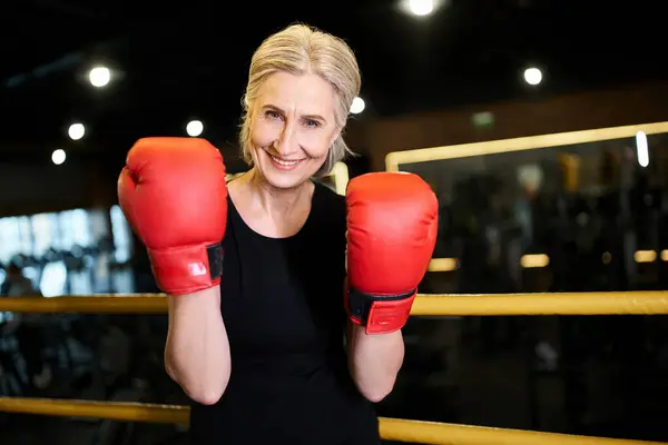 Pretty cheerful athletic senior woman with boxing gloves smiling at camera while on ring in gym — Stock Photo