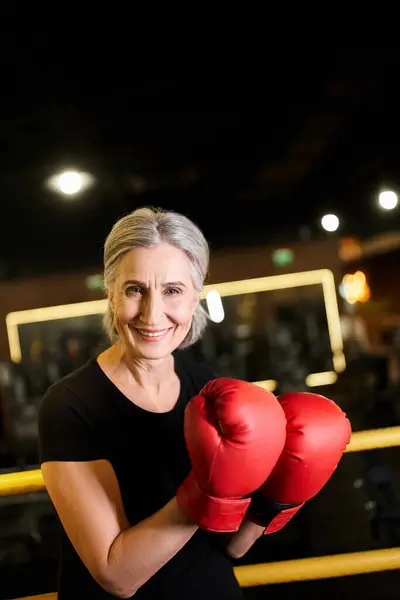 Good looking cheerful senior sportswoman with boxing gloves smiling at camera while on ring in gym — Stock Photo