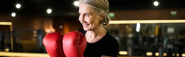 Bella donna anziana allegra in abbigliamento sportivo con i capelli grigi e guanti da boxe in posa sul ring, banner — Foto stock