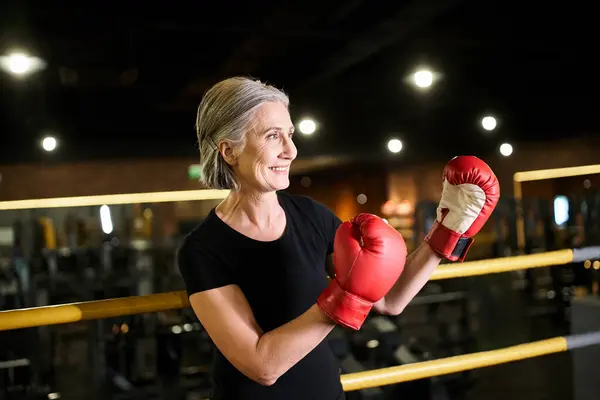Beautiful athletic senior sportswoman with gray hair and boxing gloves posing on ring in gym — Stock Photo