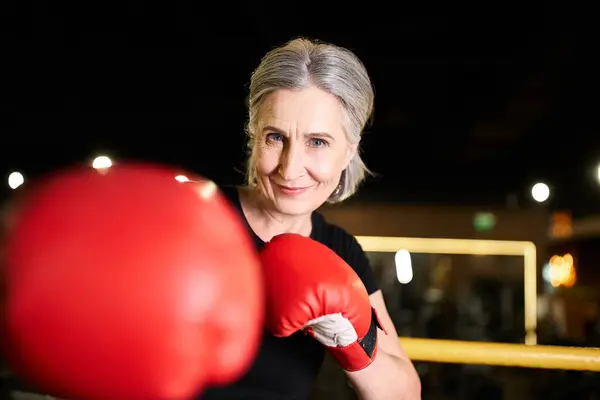 Довольно позитивная старшая спортсменка с боксерскими перчатками тренирующаяся на ринге и улыбающаяся перед камерой в тренажерном зале — стоковое фото