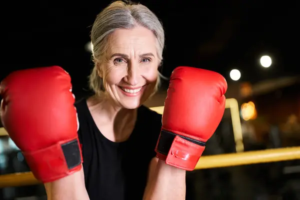 Привлекательная веселая пожилая женщина в спортивной одежде в боксёрских перчатках, тренирующаяся на ринге и улыбающаяся в камеру — стоковое фото