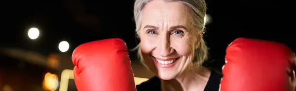 Mujer madura alegre en ropa deportiva con guantes de boxeo de entrenamiento en el anillo y mirando a la cámara, pancarta — Stock Photo