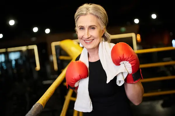 Красивая, радостная, зрелая спортсменка с полотенцами и боксерскими перчатками, улыбающаяся в камеру на ринге — стоковое фото
