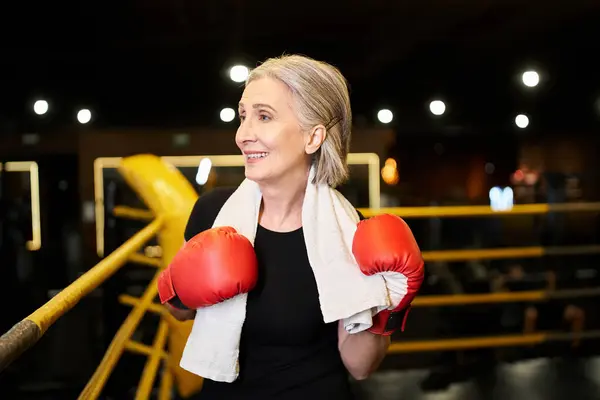 Femme âgée aux cheveux gris athlétique avec serviette et gants de boxe regardant loin sur l'anneau dans la salle de gym — Photo de stock