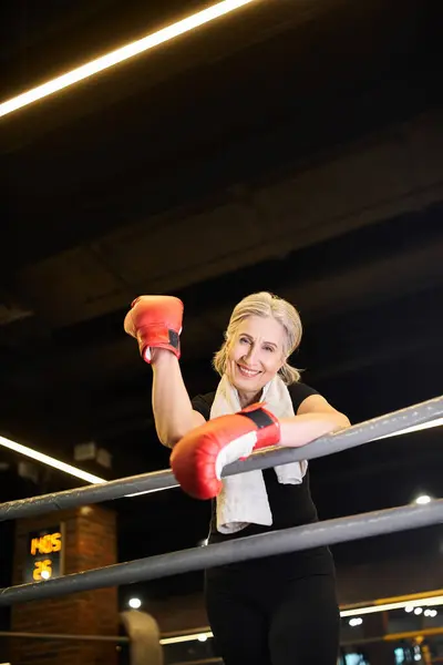 Радостная красивая зрелая женщина с полотенцами и боксерскими перчатками, тренирующаяся на ринге и улыбающаяся перед камерой — стоковое фото