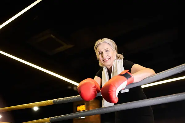 Allegra donna anziana sportiva con asciugamano e guanti da boxe che si esercitano sul ring e sorridono alla macchina fotografica — Foto stock